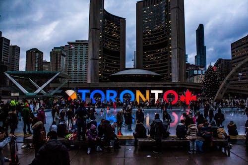 Crowd gathered at the illuminated TORONTO sign at Nathan Phillips Square in Toronto during the evening, with people ice skating in front of Toronto City Hall and city skyscrapers in the background.